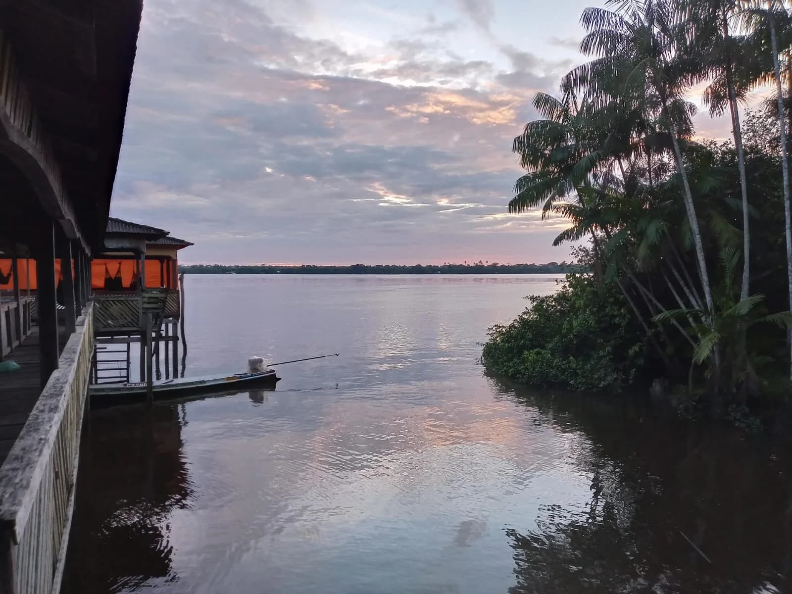 Fim de tarde no rio, visto da varanda de uma casa de palafita: uma canoa amarrada, açaizeiros na margem e o céu rosado refletido na água.