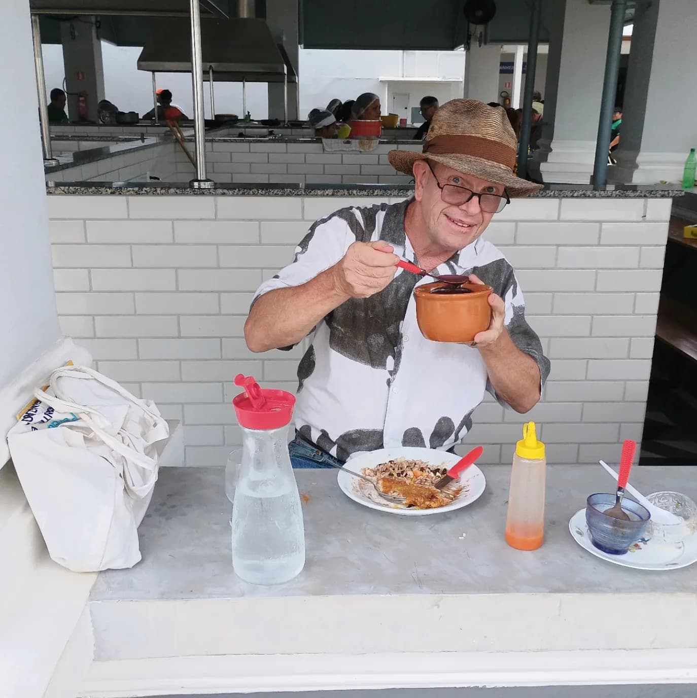 Marc toma açaí numa banca de mercado, de chapéu de palha e sorrindo para a câmara.