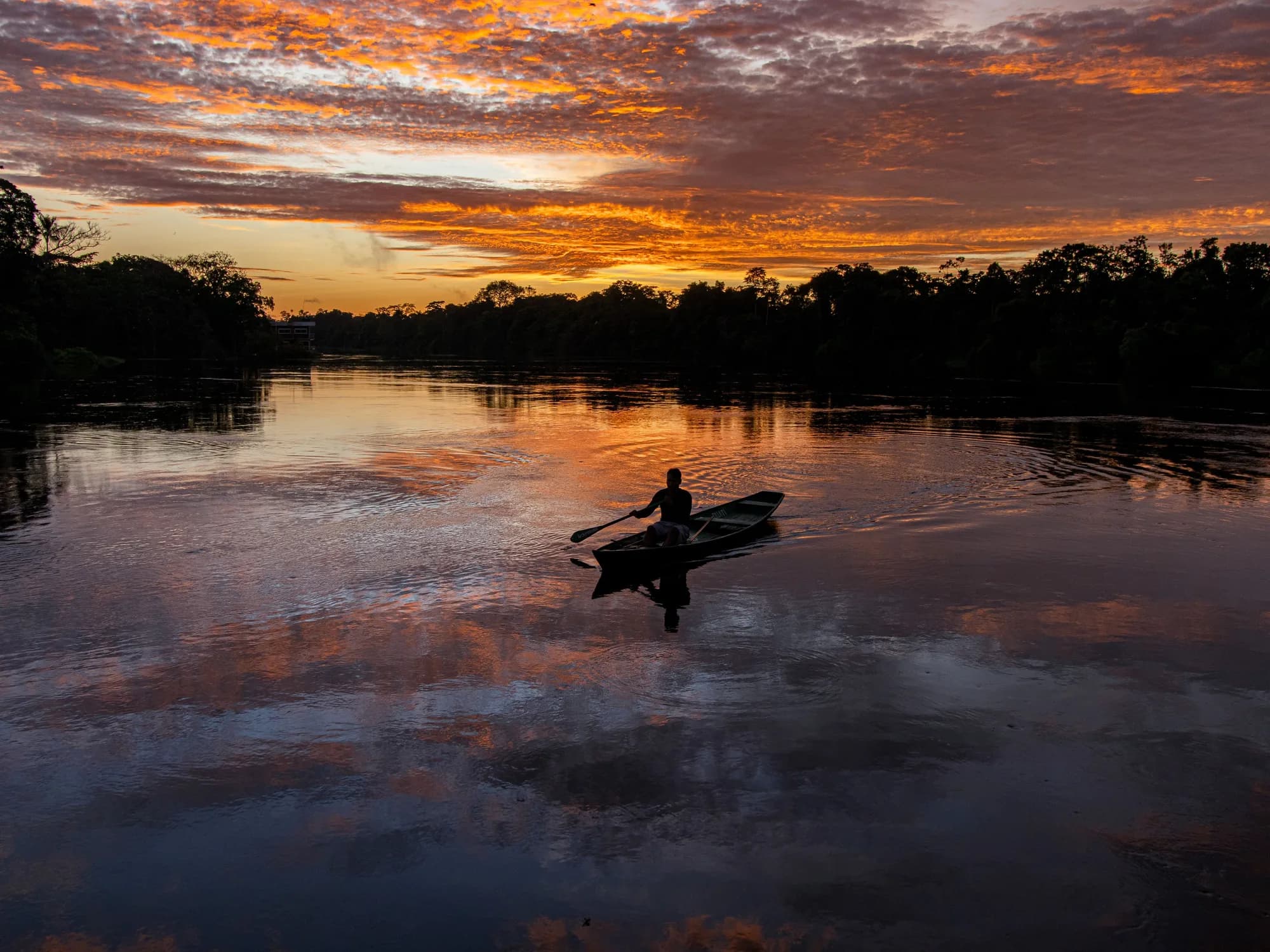 Um homem rema uma canoa num rio amazónico ao entardecer, contra o céu alaranjado e a mata na outra margem.